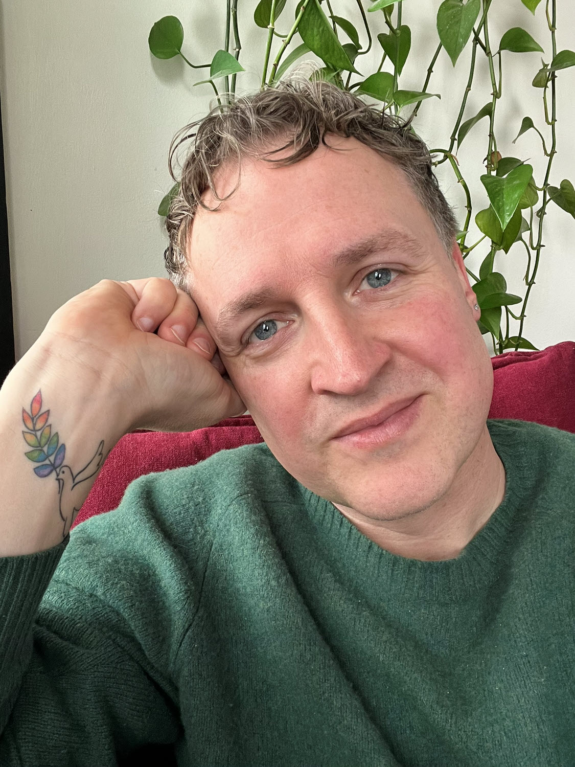 Stephen Rathjen, FIMBY's founder, wearing a green sweater, with a tattoo of a dove carrying a rainbow olive branch on his wrist